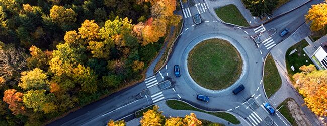 Luftaufnahme eines Kreisverkehrs mit Zebrastreifen, umgeben von herbstlich gefärbten Bäumen in gelb, orange und grün.