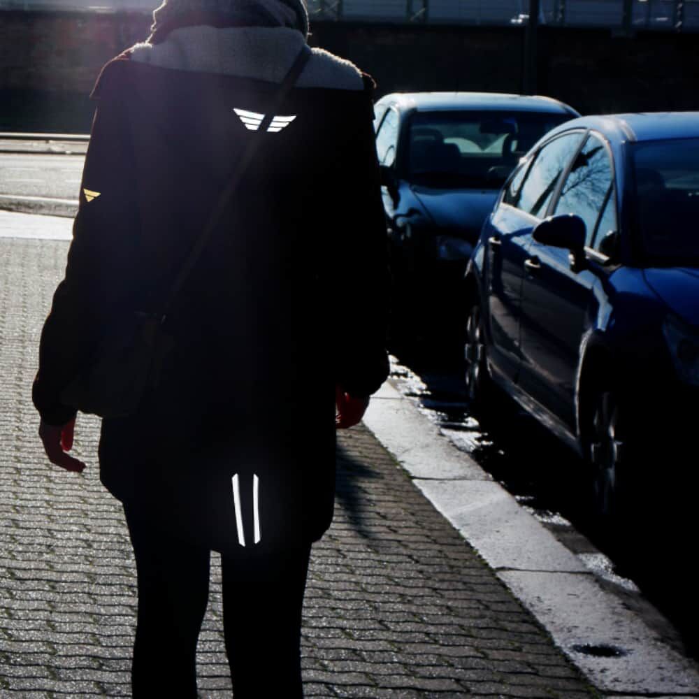 Fußgängerin mit Mantel; Reflexstreifen leuchten rückseitig im Straßenverkehr.