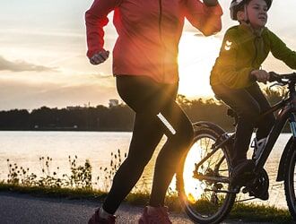 Das Bild zeigt eine Person beim Joggen neben einem Kind auf dem Fahrrad bei Sonnenuntergang am Wasser.