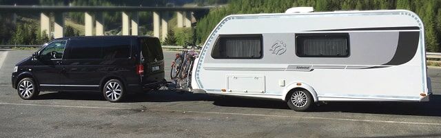 Ein Van mit einem konturmarkierten Wohnwagen im Schlepptau fährt durch eine idyllische Landschaft.
