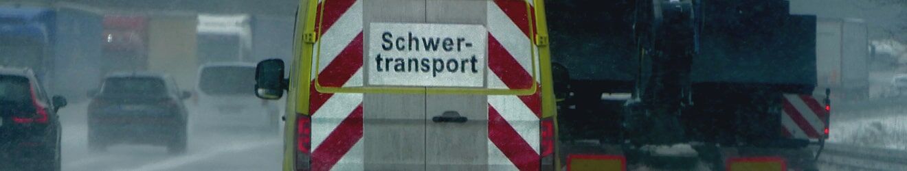 Begleitfahrzeug mit "Schwertransport"-Schild und Warnmarkierungen bei Regenwetter im Straßenverkehr.