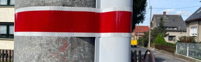 Laternenring VZ 394 – Kennzeichnung von nicht permanenter nächtlicher Straßenbeleuchtung Ein rot-weißes Reflexband um einen grauen Pfosten gewickelt.