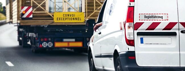Das Bild zeigt ein weißes Begleitfahrzeug mit rot-weißen Warnstreifen hinter einem Schwertransport mit gelbem "CONVOI EXCEPTIONNEL"-Schild.