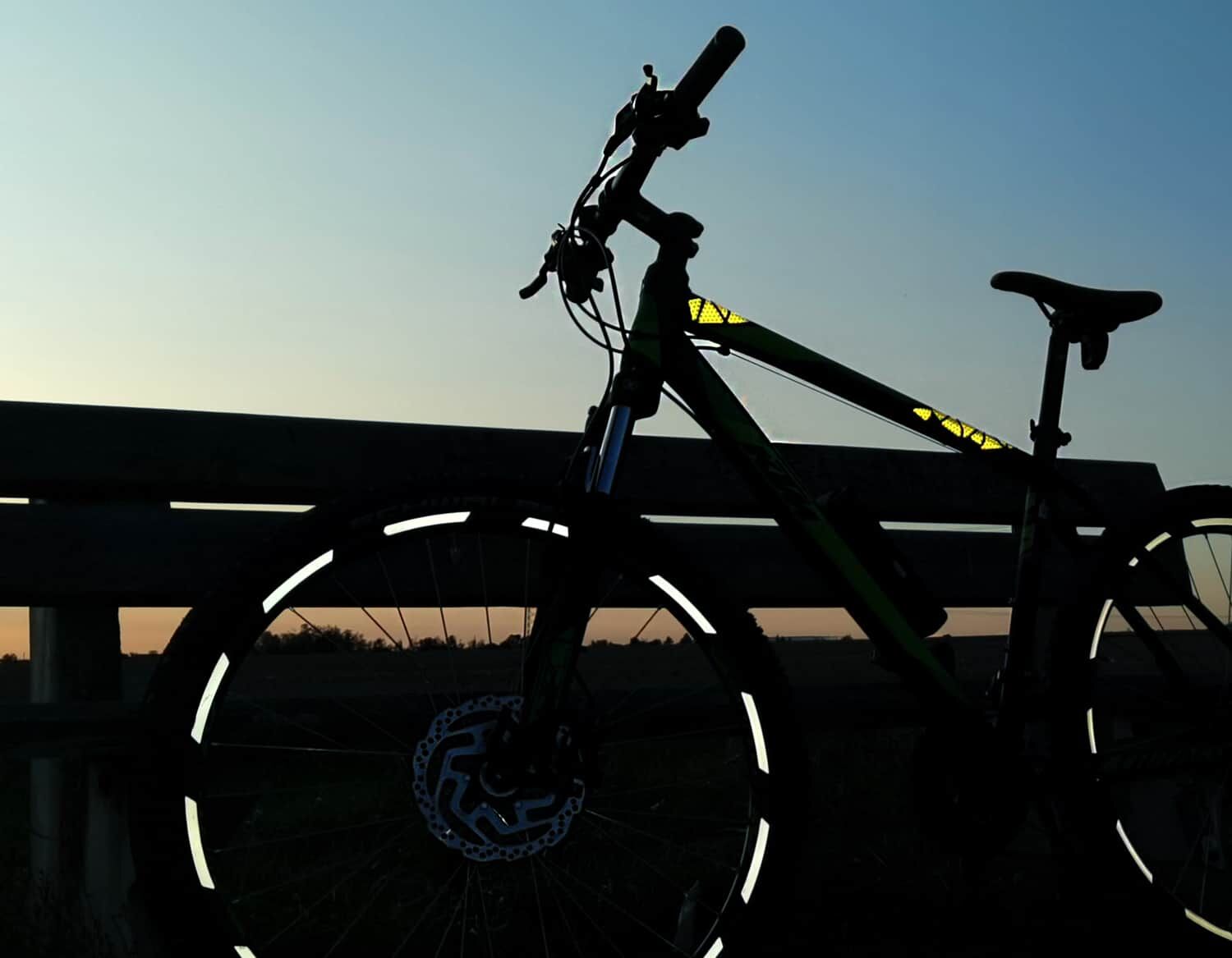 reflexsticker – Sichtbar und sicher durch den Alltag! Das Bild zeigt ein Mountainbike mit reflektierenden Felgen und Rahmen vor einer Bank bei Dämmerung, Strommasten am Horizont.