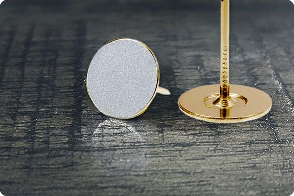 Eine Nahaufnahme zweier runder Reflektorpins: ein weißer liegend und ein goldener stehend auf dunkler Holzoberfläche.