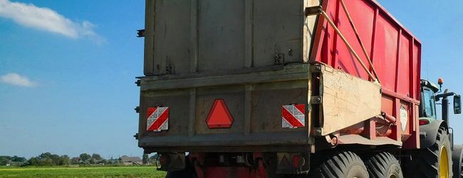 Rote Landwirtschaftsanhänger mit Warndreieck und Reflektoren, an Traktor gekoppelt bei blauem Himmel.