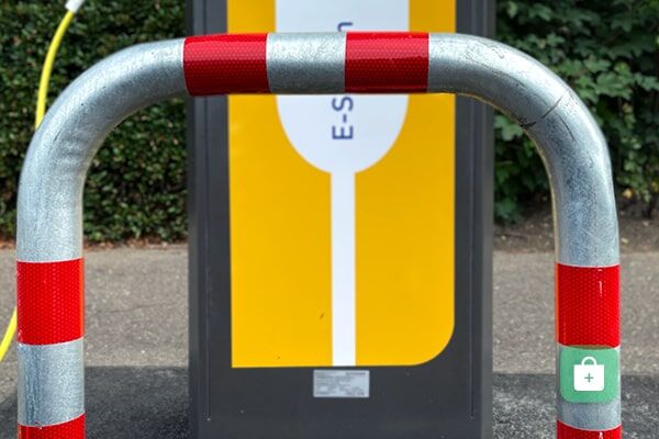 Ein Ladesäulen Rammschutz-Bügel mit roten Reflexbändern vor einer E-Ladestation und grünem Hintergrund.