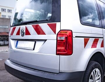 Das Bild zeigt die Rückseite eines weißen VW-Transporters mit rot-weißen Warnmarkierungen und roten Rücklichtern.