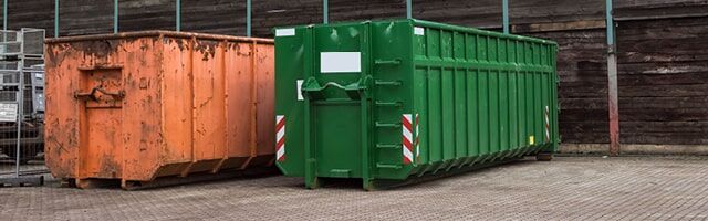 Containerwarnmarkierung gemäß § 32 StVO Ein oranger und ein grüner Abrollcontainer mit rot-weißen Warnmarkierungen an den Ecken, stehend vor einer Holzwand.