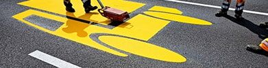 Aufbringen einer gelben Fahrbahnmarkierung mit Schablone auf frischem Asphalt.