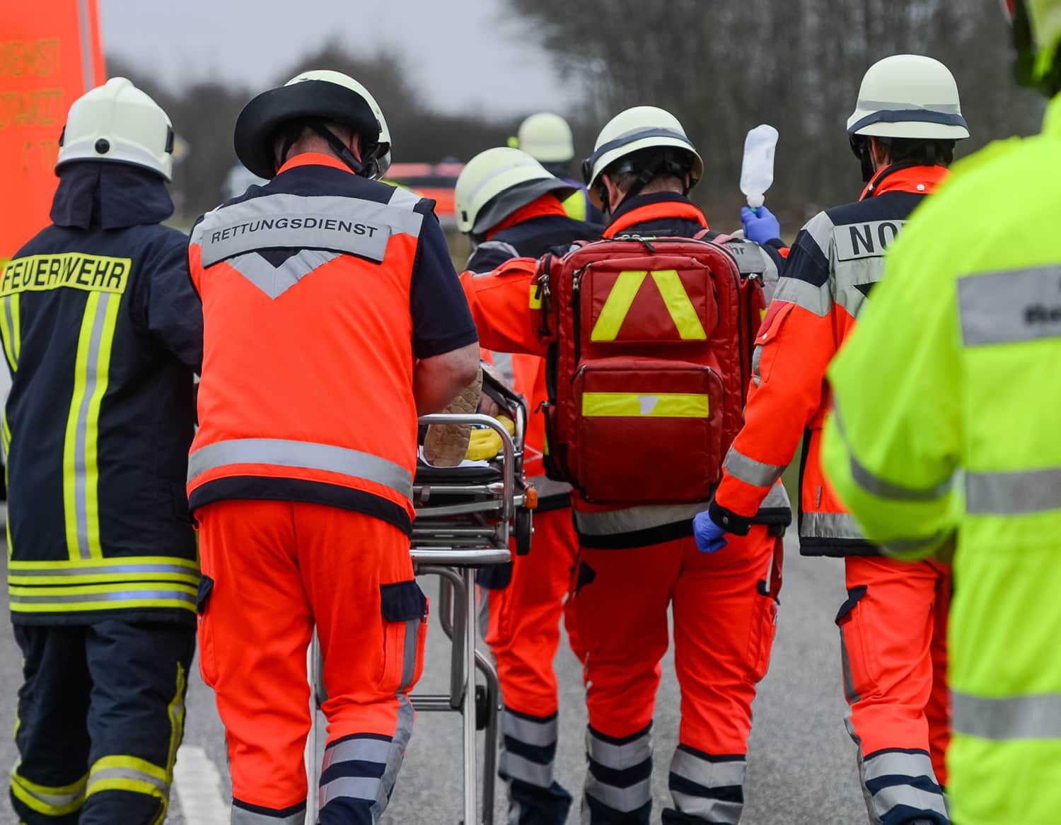 Rettungskräfte in verschiedenfarbigen Warnwesten (orange, gelb, rot) transportieren eine Trage bei einem Einsatz.