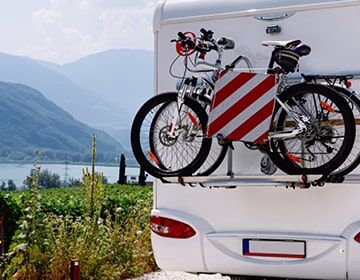 Das Bild zeigt ein weißes Wohnmobil mit Fahrrädern und rot-weißen Warnmarkierungen vor einer Berglandschaft mit See.