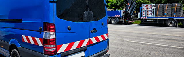 Kfz-Warnmarkierung gemäß DIN 30710 Ein blauer Transporter mit rot-weißer Warnmarkierung an Heck und Seiten beklebt ist im Anschnitt zu sehen, wie er auf einer Autbahnbasutelle abgestellt ist.