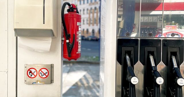 Eine Tankstelle mit rotem Feuerlöscher, Verbotsschildern (Feuer- und Rauchverbot) und Zapfsäulen im Hintergrund.