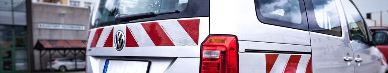 Das Bild zeigt die Rückseite eines weißen VW-Transporters mit rot-weißen Warnmarkierungen und roten Rücklichtern.