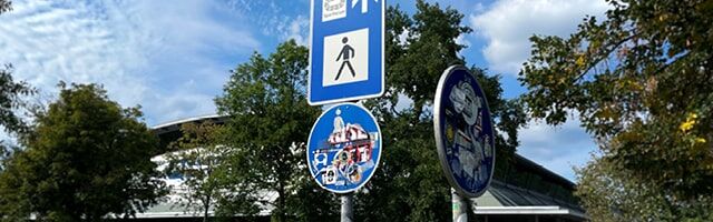 ASTIFOL® Anti Sticker Folie ORALITE® 5097 – Allrounder Schutzschicht für Verkehrszeichen, Hinweisschilder & Co. Ein mit Astifol geschütztes Verkehrsschild sticht beschadigungsfrei zwischen zwei kaum erkennbaren, vollgestickerten Schildern vor einem Gebäude im Hintergrund heraus.