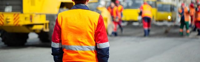 Person in orangener Warnweste von hinten, im Hintergrund weitere Arbeiter und gelbe Fahrzeuge