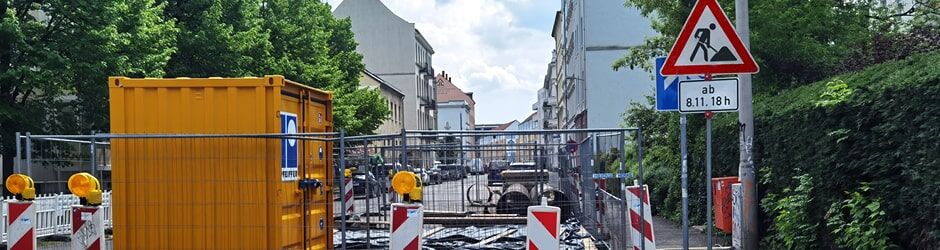 Eine Baustelle mit gelbem Container, Absperrungen und Warnleuchten in einer städtischen Wohnstraße.