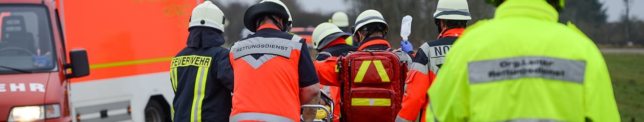 Rettungskräfte in verschiedenfarbigen Warnwesten (orange, gelb, rot) transportieren eine Trage bei einem Einsatz.