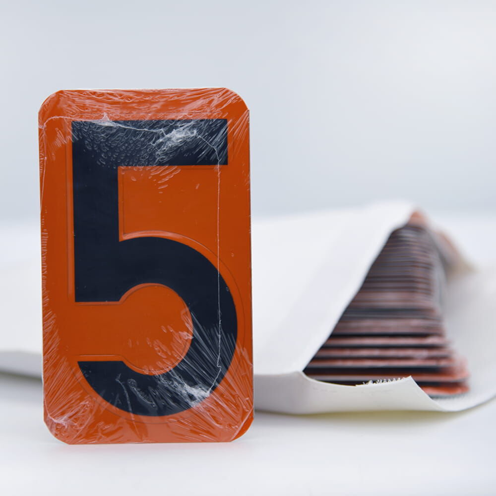 ADR-Zifferntafel mit schwarzer 5 in Schutzfolie, stehend vor weißem Umschlag mit weiteren Tafeln.