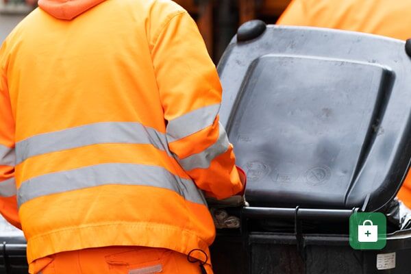 Ein Arbeiter der Müllabfuhr in orangefarbener Warnschutzjacke mit reflektierenden Streifen neben einer grauen Mülltonne.