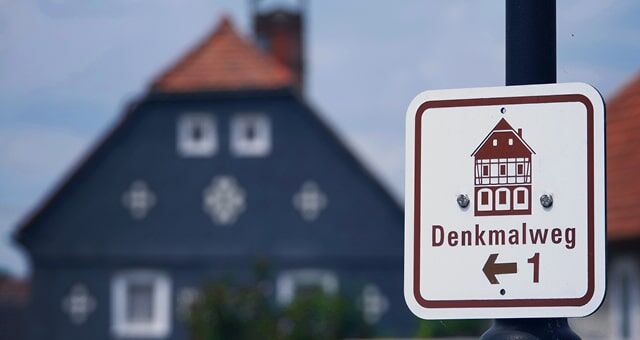 Schild „Denkmalweg“ vor traditionellem Fachwerkhaus im Hintergrund