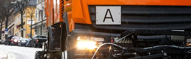 Ein orangefarbenes Nutzfahrzeug mit eingeschaltetem Licht und einer Abfall-Warntafel an der Seite.