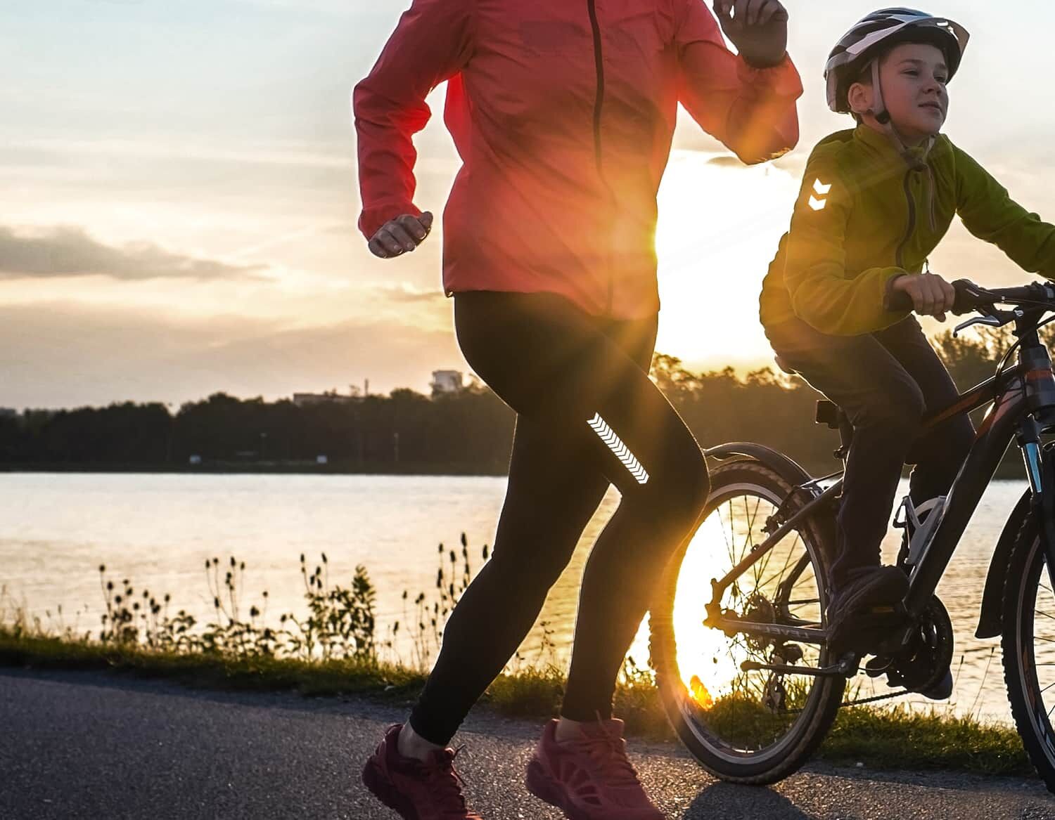 Das Bild zeigt eine Person beim Joggen neben einem Kind auf dem Fahrrad bei Sonnenuntergang am Wasser.