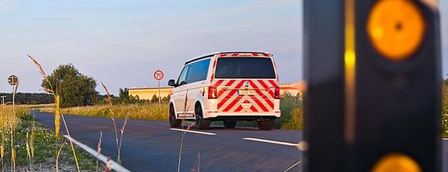 Ein Einsatzfahrzeug mit rot-weißer Warnmarkierung fährt auf einer Landstraße mit Geschwindigkeitsmessgerät im Vordergrund.
