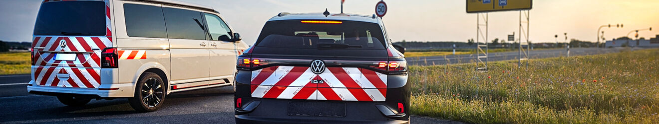 Rückansicht von zwei Fahrzeugen mit rot-weißen Warnmarkierungen - ein schwarzer VW und ein weißer VW-Transporter auf einer Straße neben einer Wiese.
