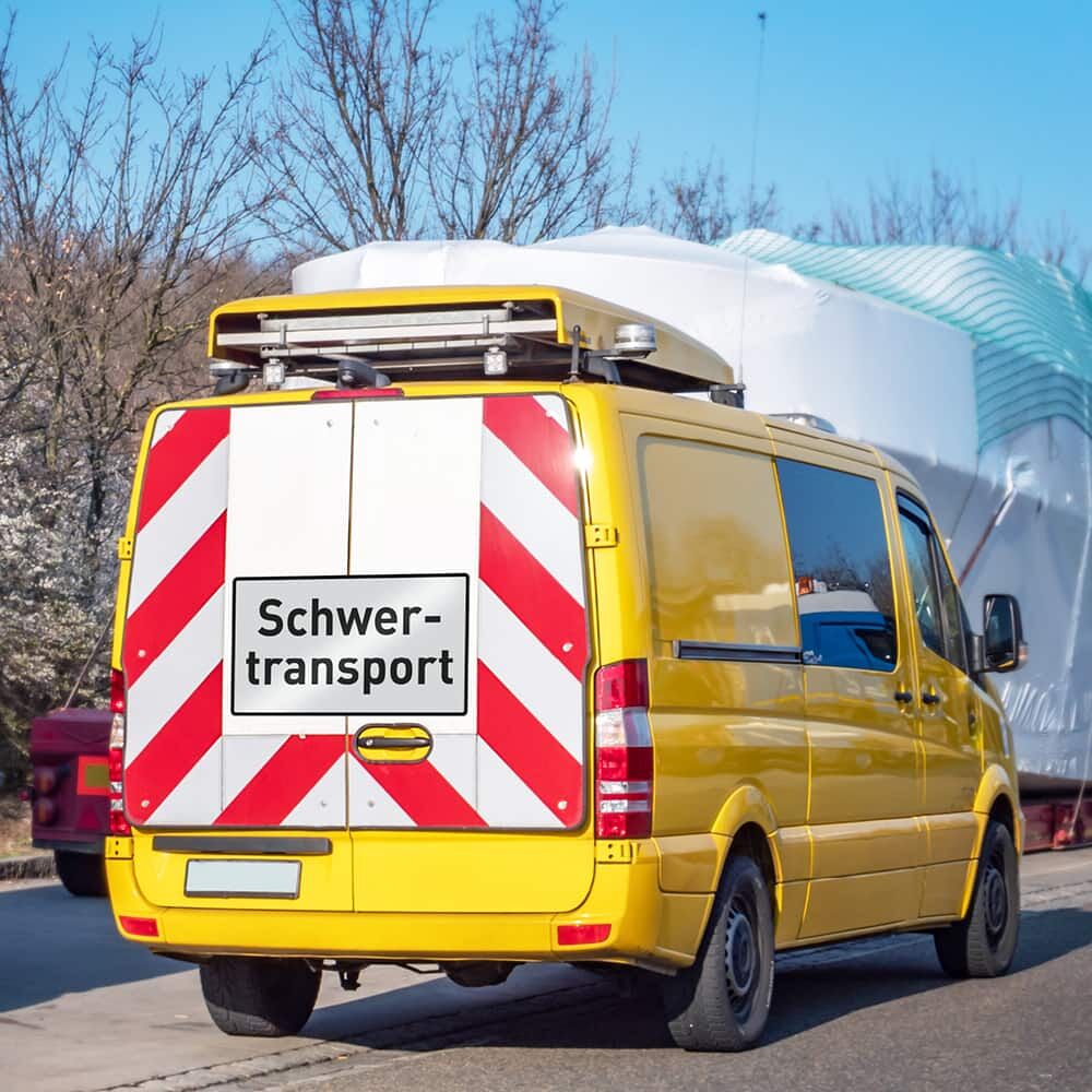Schwertransportschild am gelben Begleitfahrzeug, großes Ladegut im Hintergrund