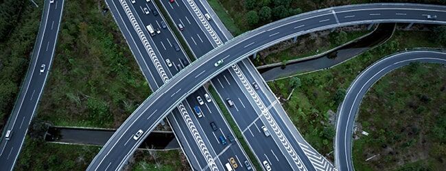 Luftaufnahme einer mehrspurigen Autobahnkreuzung mit Überführungen und fahrenden Fahrzeugen, umgeben von grünen Waldflächen.