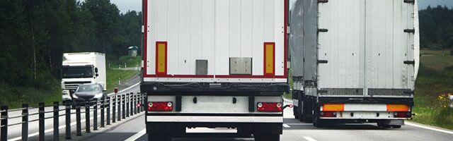 Heckwarnmarkierung nach ECE 70 Zwei Lastkraftwagen mit Heckwarnmarkierungen fahren auf einer Autobahn, umgeben von Bäumen im Hintergrund.