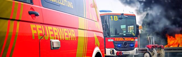 Kennzeichnung von Feuerwehrfahrzeugen nach DIN 14502-3 Mehrere Feuerwehrwagen sind vor einem in Flammen stehenden Gebäude positioniert, um den Brand zu löschen.