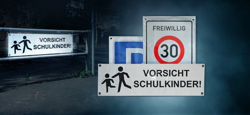 Die Schilderbanner zeigen eine Tempo-30-Zone „FREIWILLIG“ und zwei Warnschilder mit der Aufschrift „VORSICHT SCHULKINDER!“ mit Piktogrammen von einem erwachsenen Menschen und einem Kind. Die Schilder sind nachts oder bei Dunkelheit fotografiert und sind r