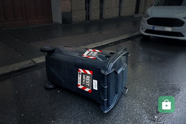 Ein umgekippter schwarzer Müllcontainer auf der regennasser Straße neben einem geparkten Auto.