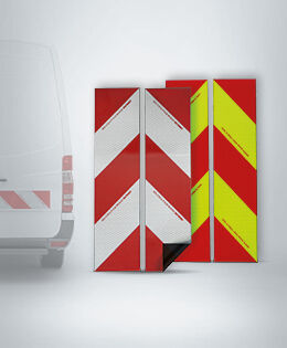 Reflektierende Warnmarkierungen in rot-weiß und rot-gelb (chevron-förmig) für Fahrzeuge. Links ist ein Teil eines weißen Transporters zu sehen.