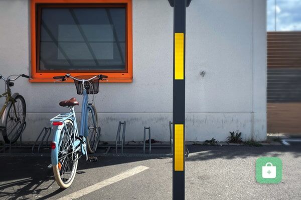 Ein schwarzer Pfosten eines Unterstandes mit gelben Reflexstreifen steht neben einem abgestellten hellblauen Fahrrad und einer grauen Hauswand.
