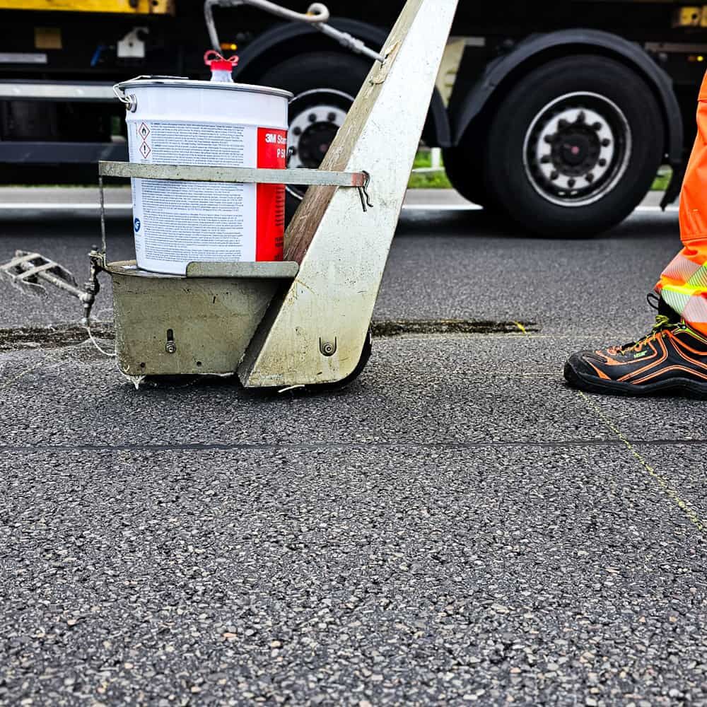 P50-Eimer im Fahrwagen auf Asphalt, Straßenbauer in Warnschuh daneben