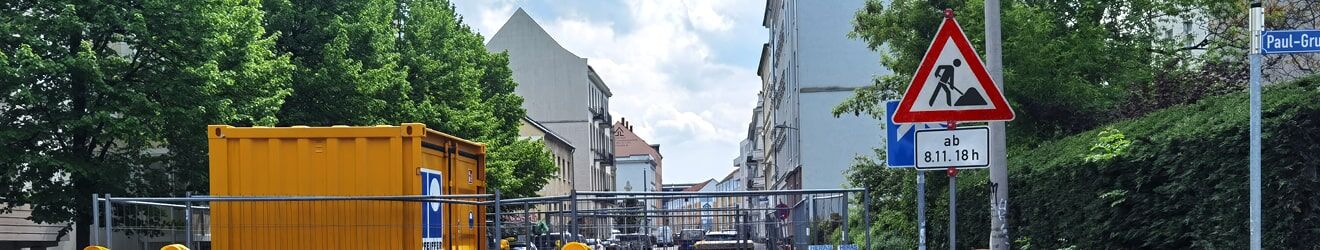 Eine Baustelle mit gelbem Container, Absperrungen und Warnleuchten in einer städtischen Wohnstraße.