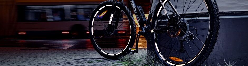 Mountainbike mit reflektierenden Felgen bei Nacht auf nasser Straße, unscharfe Lichter eines vorbeifahrenden Busses.