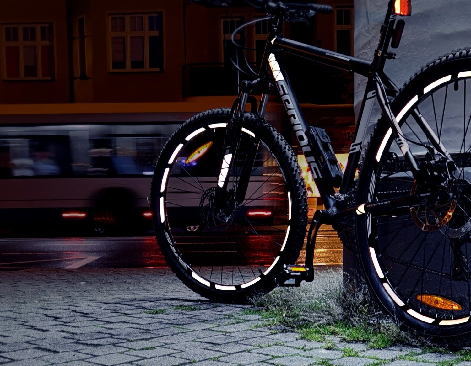 Mountainbike mit reflektierenden Felgen bei Nacht auf nasser Straße, unscharfe Lichter eines vorbeifahrenden Busses.