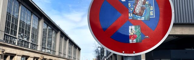 Schutzlaminate für Verkehrszeichen Verksschild Halteverbot zur einen Hälfte sauber, mit Schutzfolie beklebt und zur anderen Hälfte schutzlos und vollgestickert mit Bahnhofsparkplatz im Hintergrund.