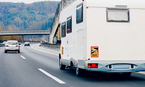 Abschnittstitelbild zu Angles Morts-Schildern Das Bild zeigt ein Wohnmobil aus Heckperspektive mit Angles Morts-Aufkleber auf der Autobahn mit Bergen im Hintergrund