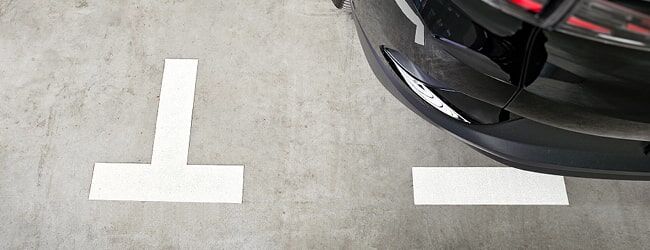 Weiße Markierungen auf einem Parkplatz mit der Ecke eines schwarzen Fahrzeugs am rechten Bildrand.