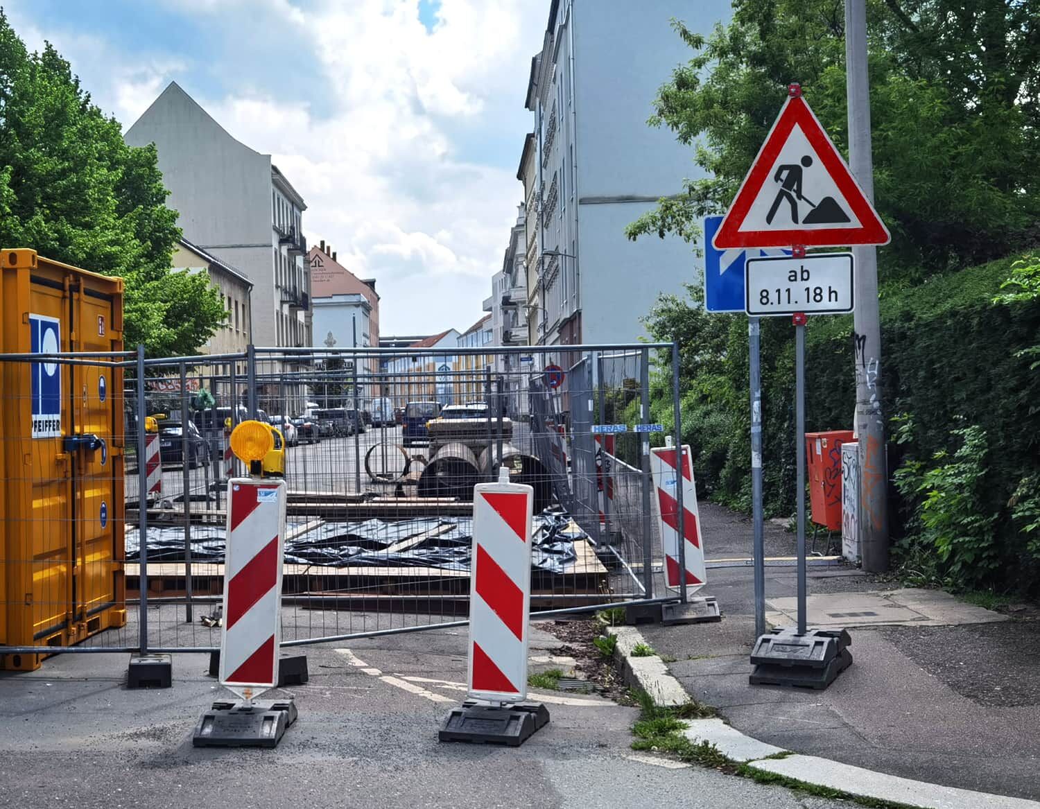 Eine Baustelle mit gelbem Container, Absperrungen und Warnleuchten in einer städtischen Wohnstraße.