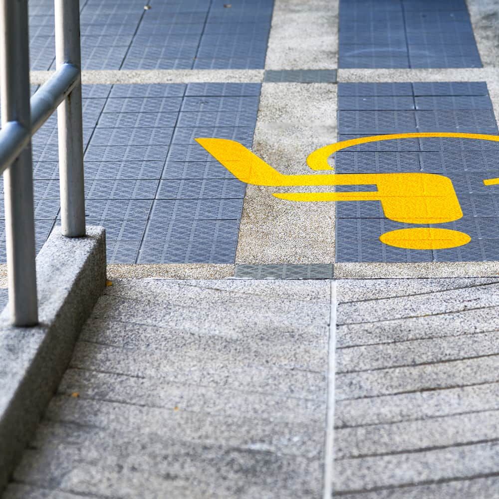 Gelbes Rollstuhlsymbol als Bodenmarkierung auf Gehweg mit Rampe und Fliesen