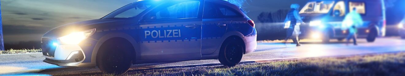 Blauer Polizeiwagen mit eingeschalteten Lichtern bei Nacht, einem weiteren Einsatzfahrzeug und Personen im Hintergrund.