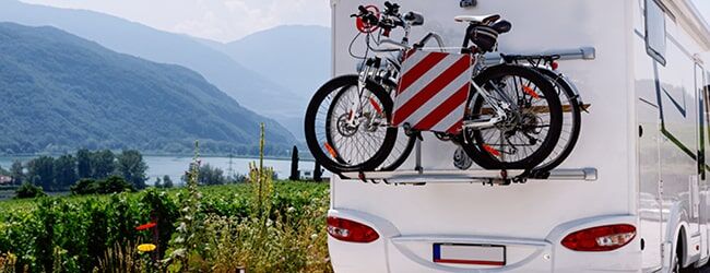 Das Bild zeigt ein weißes Wohnmobil mit Fahrrädern und rot-weißen Warnmarkierungen vor einer Berglandschaft mit See.