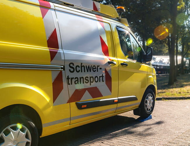 Gelbes Begleitfahrzeug mit Warnmarkierung und Schriftzug „Schwertransport“, Seitenansicht.